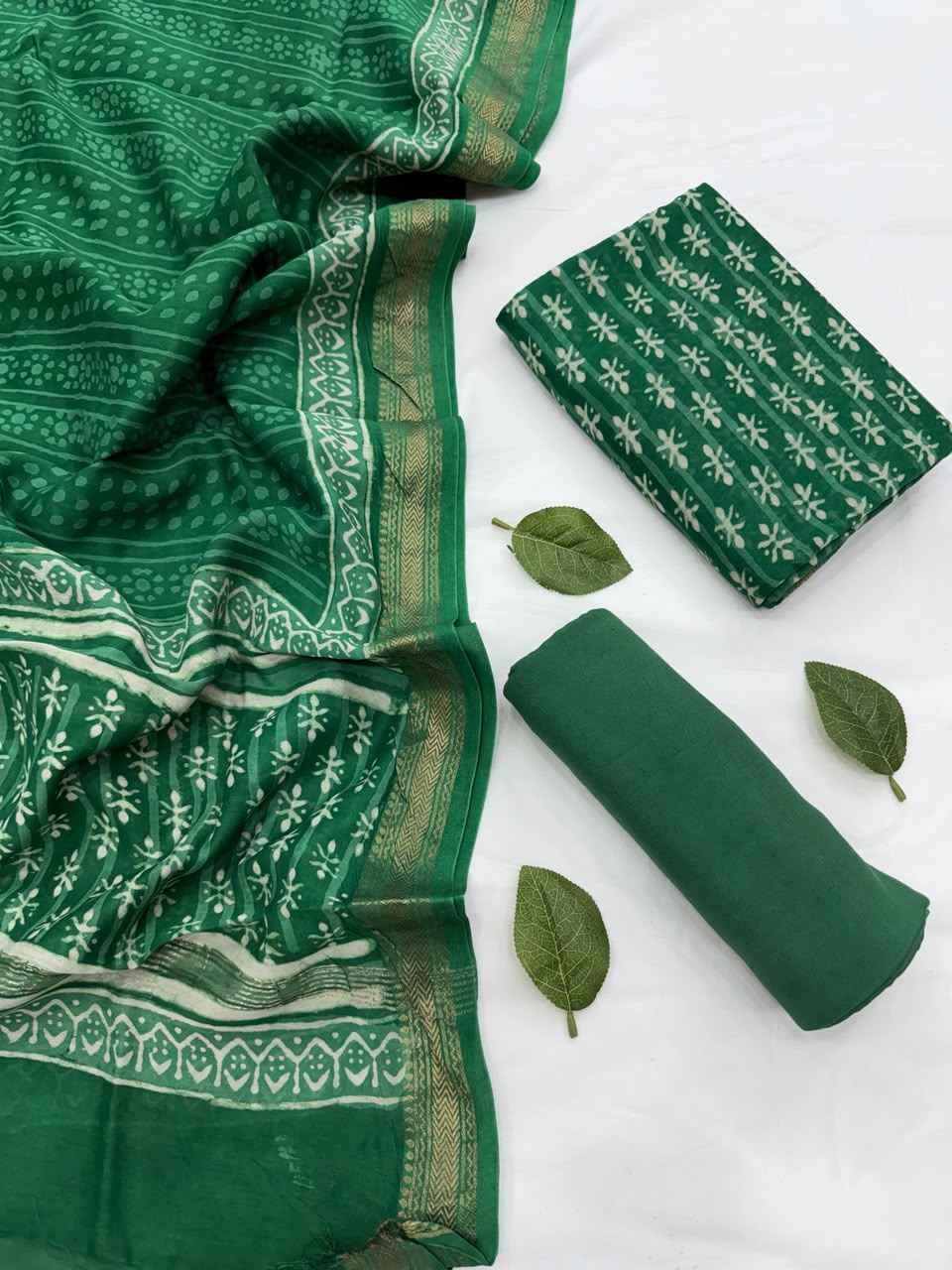 Full flat lay view of the Green Dabu Hand Block Print Maheshwari Silk Suit Set, showcasing the green kurta fabric with white patterns, a matching patterned dupatta with a golden handloom woven Zari border, and a neatly rolled solid green bottom fabric decorated with fresh green leaves.