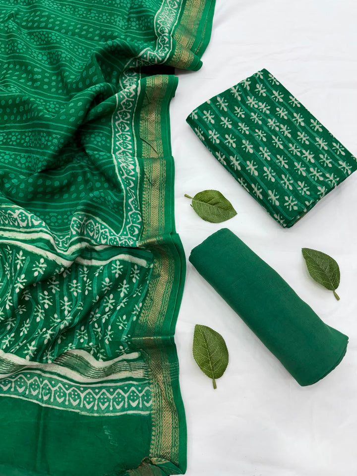 Full flat lay view of the Green Dabu Hand Block Print Maheshwari Silk Suit Set, showcasing the green kurta fabric with white patterns, a matching patterned dupatta with a golden handloom woven Zari border, and a neatly rolled solid green bottom fabric decorated with fresh green leaves.