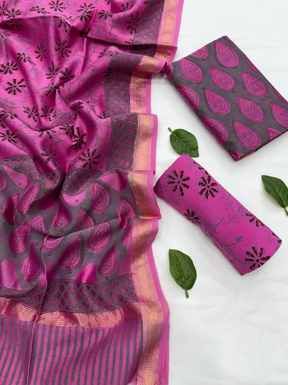 Full flat lay view of the Gulabi Pink Hand Block Print Maheshwari Silk Suit Set, displaying the charcoal grey patterned kurta fabric, a matching vibrant pink block-printed dupatta with a golden Zari border, and a neatly folded pink floral bottom fabric decorated with fresh green leaves.