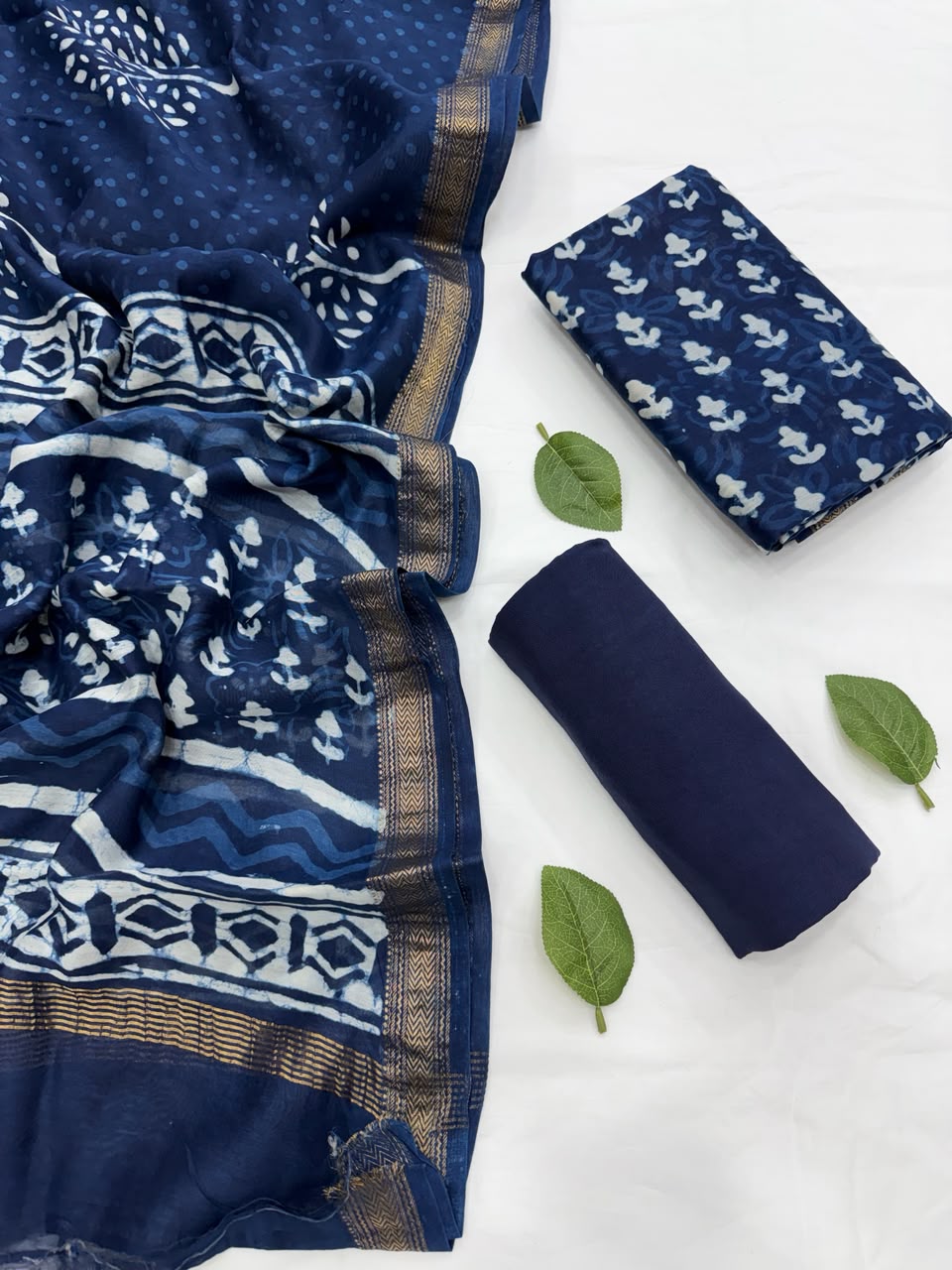 Full flat lay view of the Indigo Dabu Buti Hand Block Print Maheshwari Silk Suit Set, displaying the deep indigo kurta fabric with small scattered floral butis, a matching patterned dupatta with a golden Zari border, and a neatly rolled solid blue bottom fabric styled with fresh green leaves.