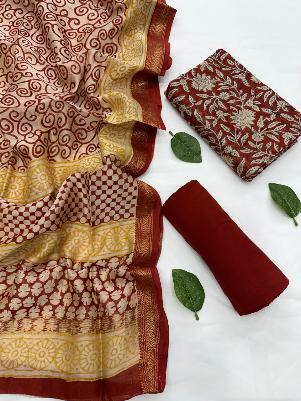 Full flat lay view of the Maroon Bagru Hand Block Print Maheshwari Silk Suit Set, featuring a cream and red patterned dupatta with a golden Zari border, a matching maroon floral kurta fabric, and a neatly rolled solid deep red bottom fabric styled with fresh green leaves.