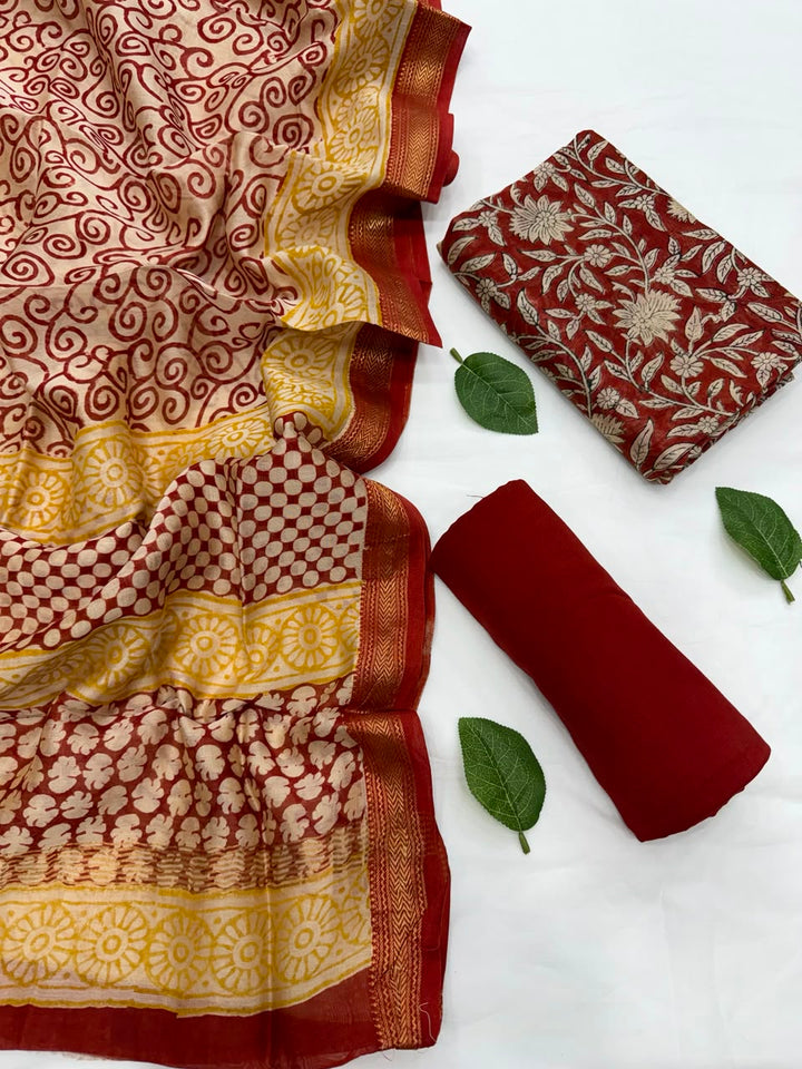Full flat lay view of the Maroon Bagru Hand Block Print Maheshwari Silk Suit Set, featuring a cream and red patterned dupatta with a golden Zari border, a matching maroon floral kurta fabric, and a neatly rolled solid deep red bottom fabric styled with fresh green leaves.