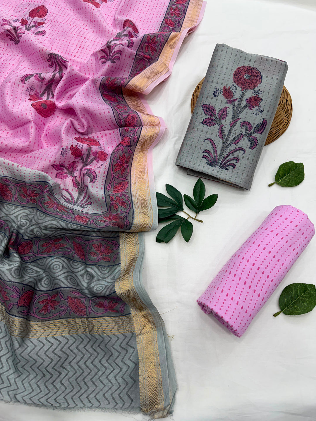 A pink and gray floral printed fabric set arranged on a white surface, including a draped dupatta, a folded gray fabric piece, and a rolled pink fabric piece, with green leaves as styling props.