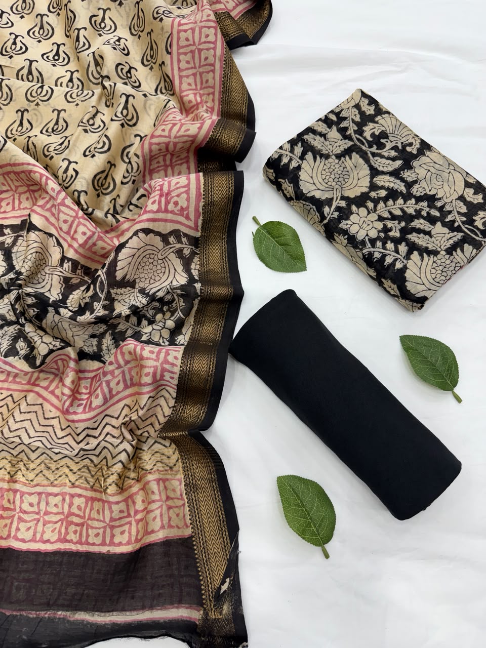 Full flat lay view of the Natural Black Bagru Print Maheshwari Silk Suit Set, displaying the black kurta fabric with golden floral motifs, a matching patterned dupatta with a golden Zari border, and a neatly rolled solid black bottom fabric styled with fresh green leaves.