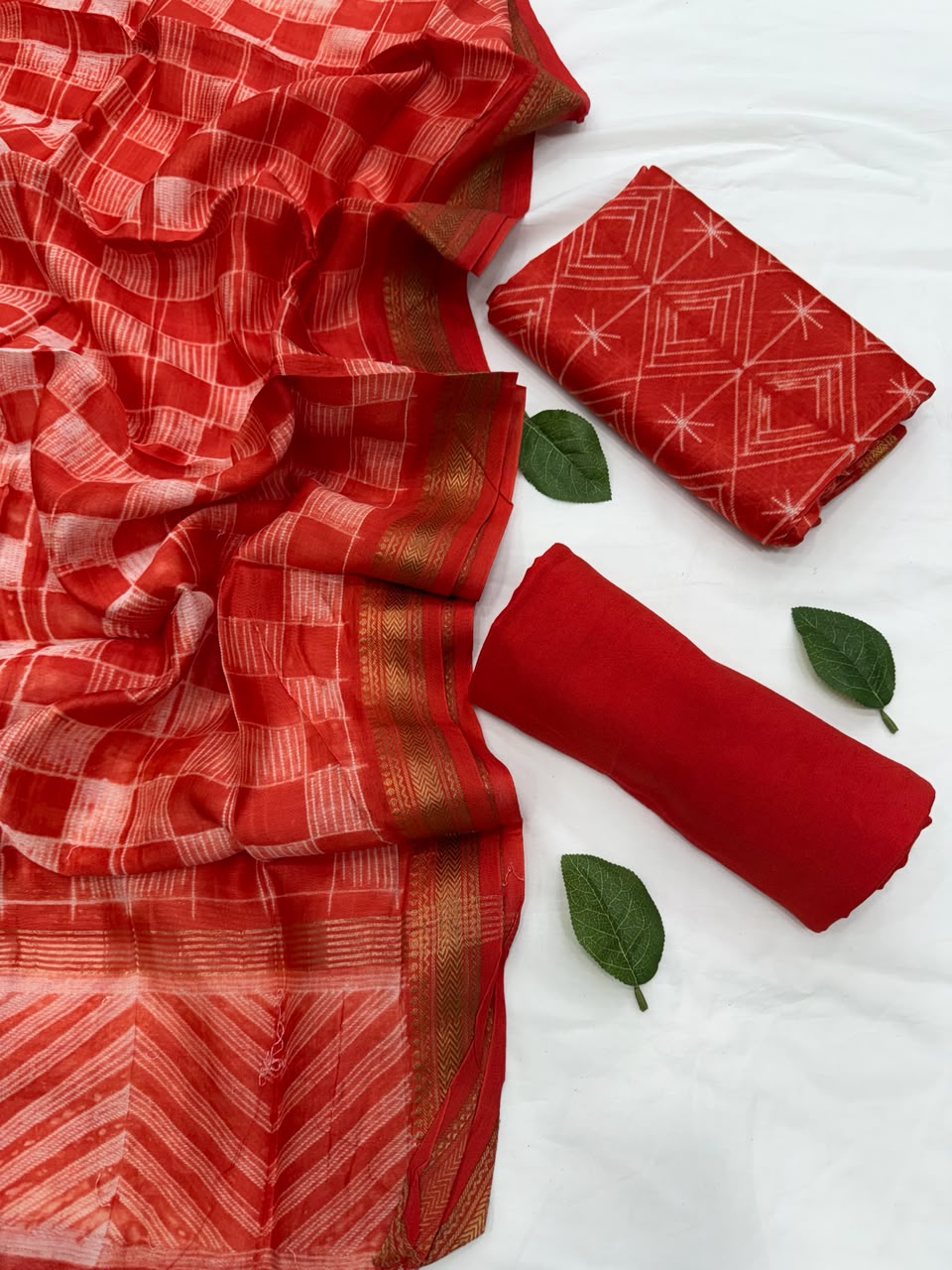 Full flat lay view of the Orange Stitching Shibori Art Maheshwari Silk Suit Set, showcasing the red-orange geometric shibori kurta fabric, matching patterned dupatta with a golden handloom woven Zari border, and a neatly rolled solid orange bottom fabric decorated with fresh green leaves.