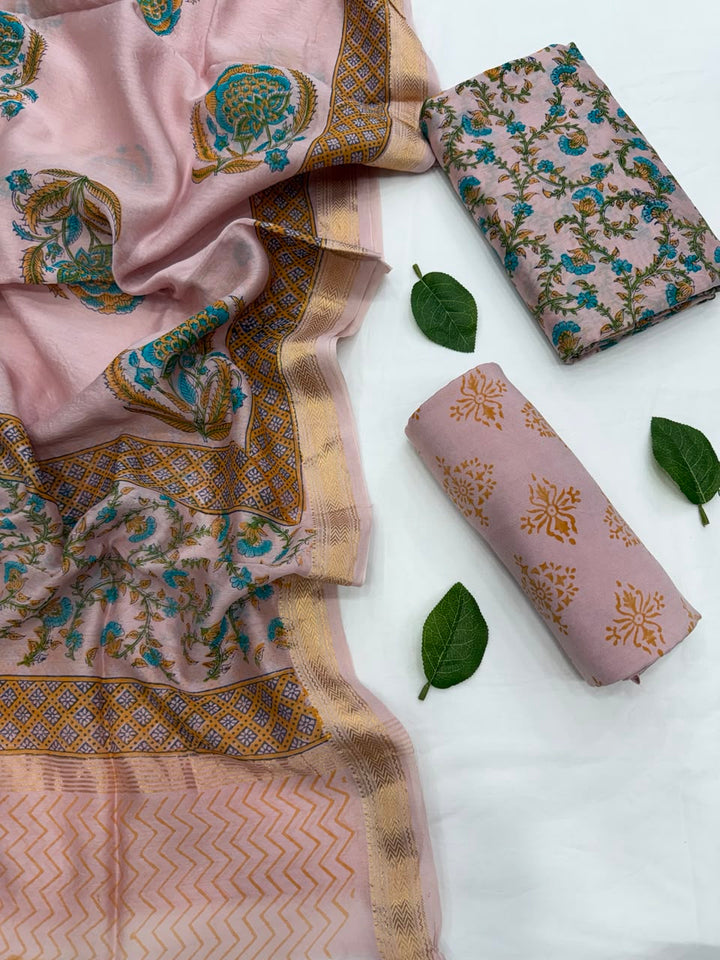 Full flat lay view of the Pink Sanganeri Block Print Maheshwari Silk Suit Set, showcasing the pink kurta fabric with blue and green floral patterns, a matching block-printed dupatta with a golden woven Zari border, and a neatly rolled solid pink bottom fabric decorated with fresh green leaves.