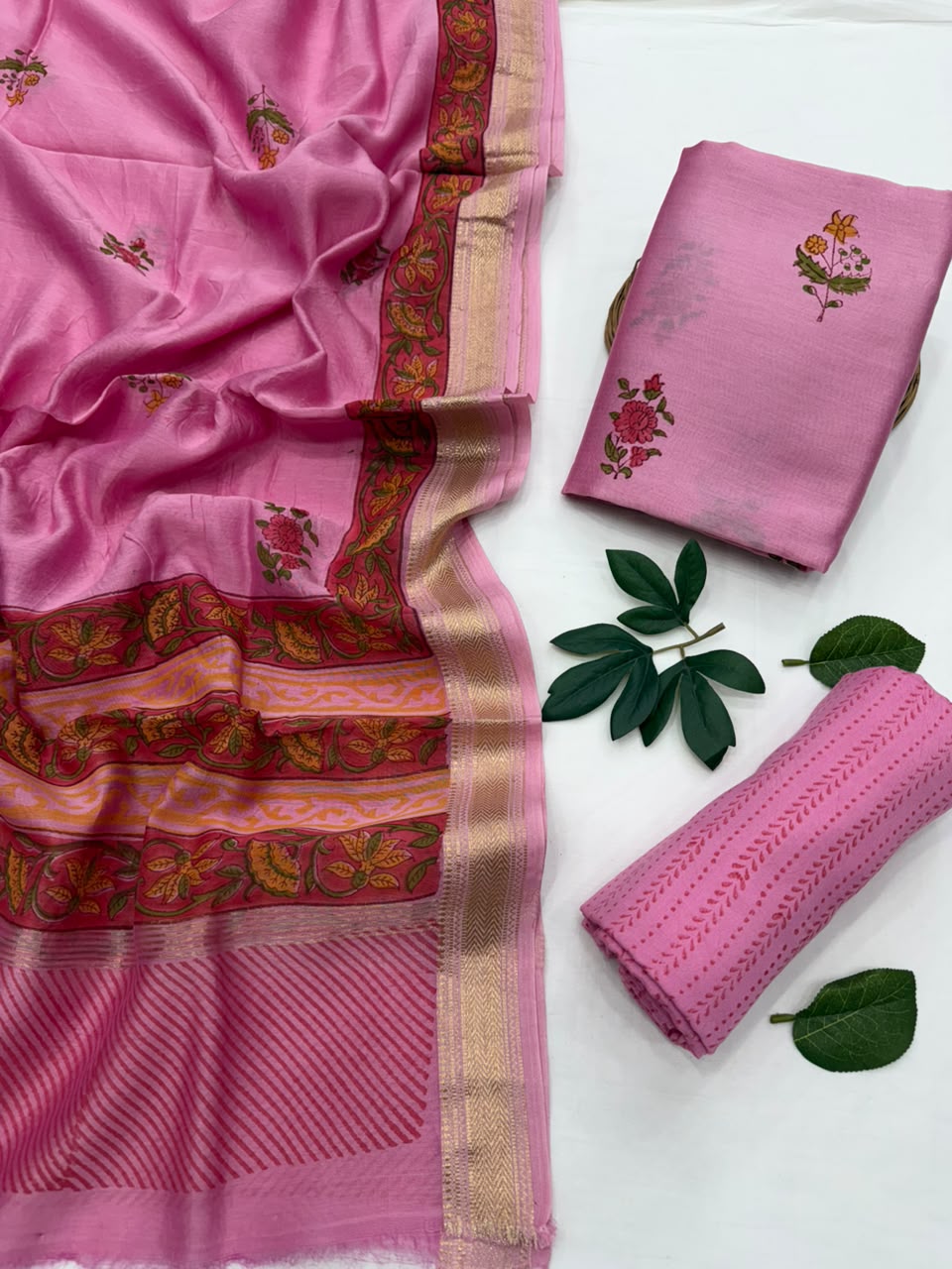 Pink silk saree with floral embroidery and a gold zari border, displayed alongside folded and rolled fabric pieces on a white surface with green leaves.