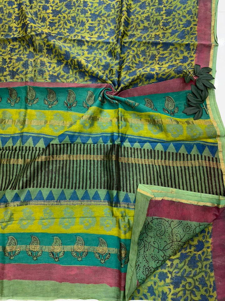 Close-up flat lay of a natural green Chanderi silk saree showcasing its intricate blue and olive botanical prints, a multi-layered geometric border, and traditional paisley motifs on the pallu.
