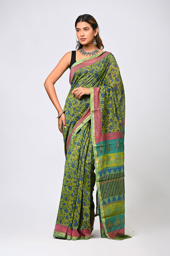 A woman modeling a natural green Chanderi silk saree featuring intricate blue and olive botanical block prints, an elegant patterned border, and a contrasting pink selvedge.