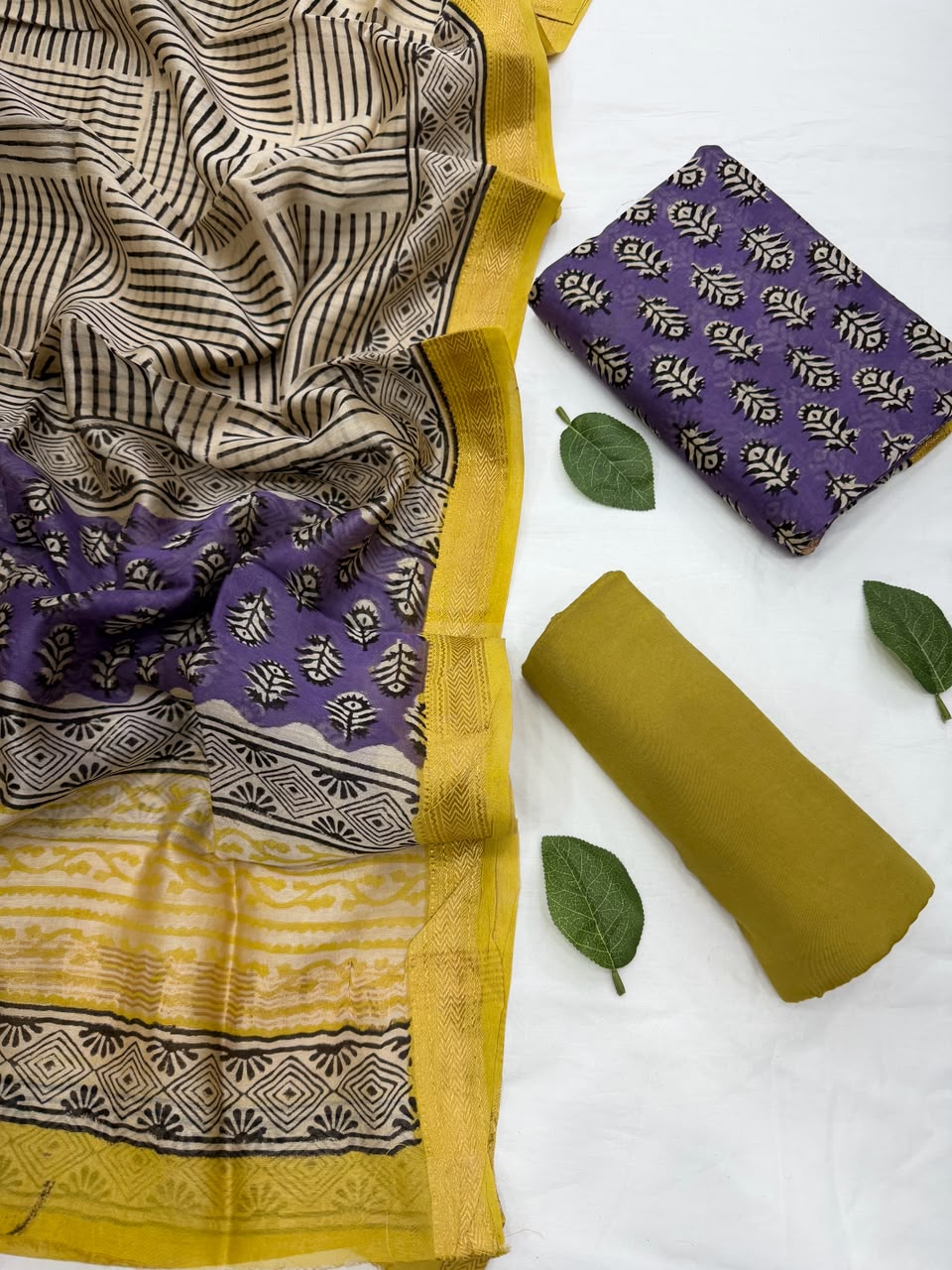 Full flat lay view of the Purple Bagru Buti Hand Block Print Maheshwari Silk Suit Set, showcasing the royal purple floral buti printed kurta fabric, matching patterned dupatta with a golden handloom Zari border, and a neatly rolled plain mustard bottom fabric styled with fresh green leaves.