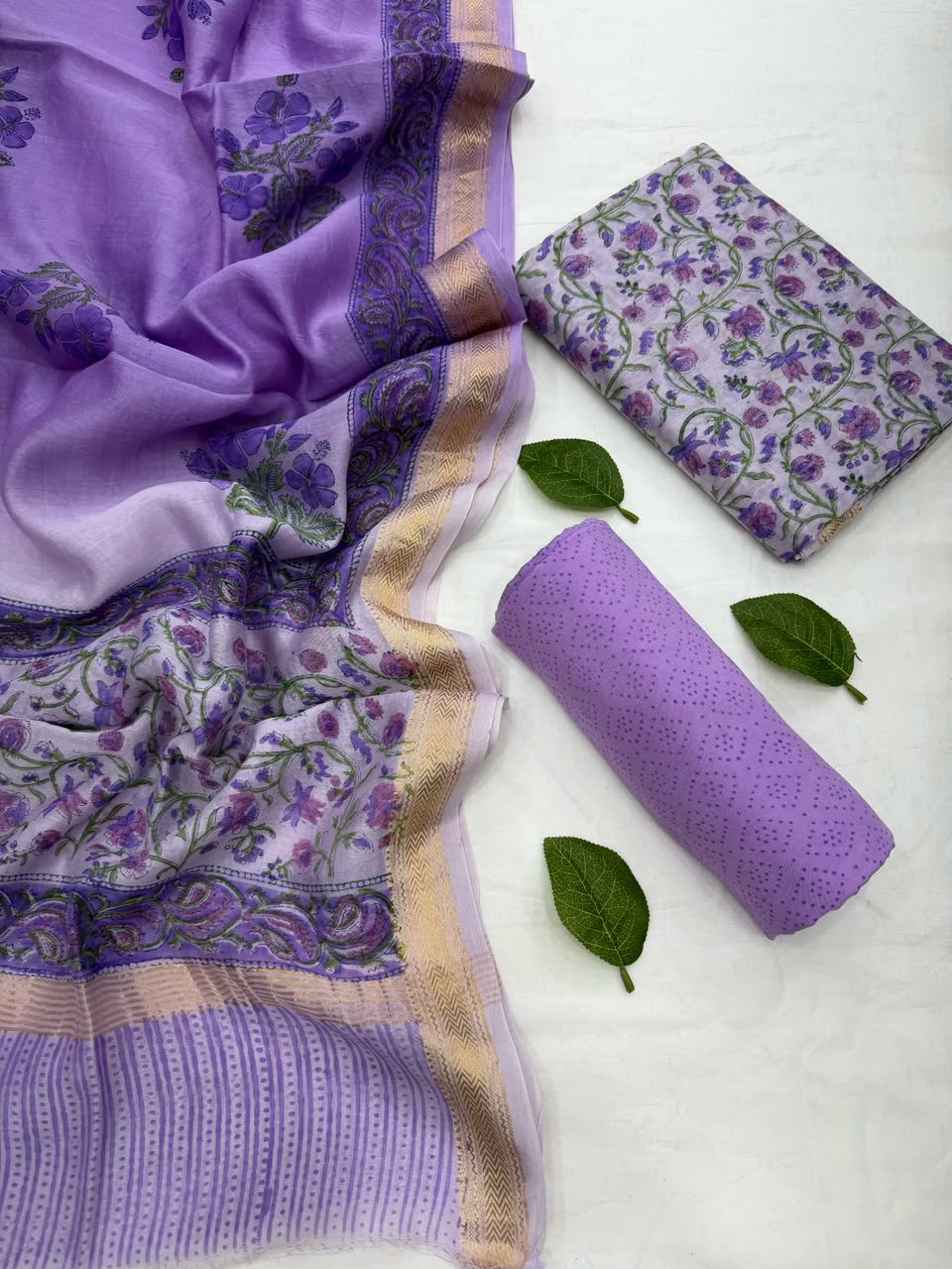 Full flat lay view of the Purple Sanganeri Jaal Print Maheshwari Silk Suit Set, showcasing the royal purple kurta fabric with intricate all-over floral lattice work, a matching block-printed dupatta with a golden handloom Zari border, and a neatly rolled solid purple bottom fabric styled with fresh green leaves.