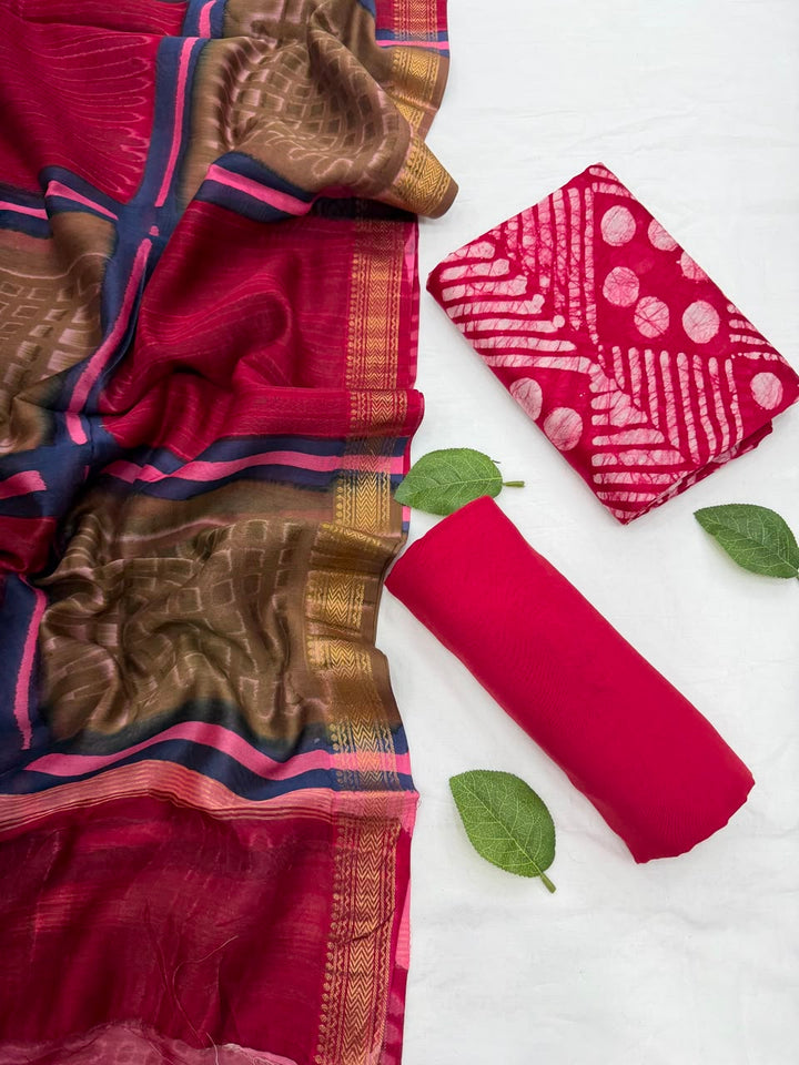 Full flat lay view of the Red Batik Print Designer Maheshwari Silk Suit Set, showcasing the red kurta fabric with white geometric batik patterns, a matching patterned dupatta with a golden woven Zari border, and a neatly rolled solid red bottom fabric decorated with fresh green leaves.
