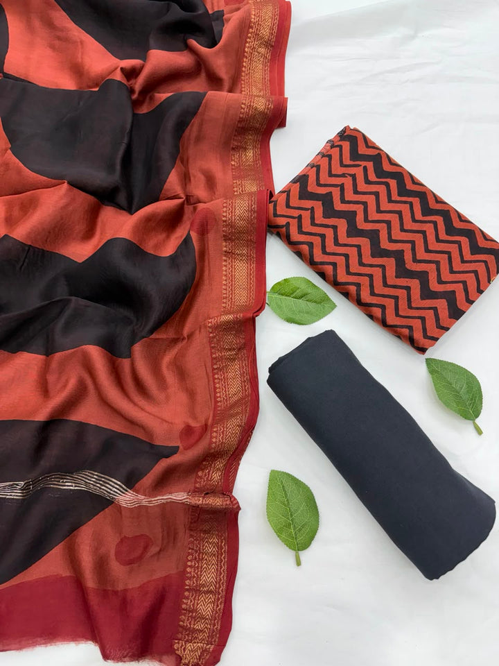 Full flat lay view of the Rust Bagru Print Designer Maheshwari Silk Suit Set, displaying the zigzag pattern kurta fabric, a solid black bottom fabric roll, and a dual-toned black and rust dupatta with a golden Zari border.
