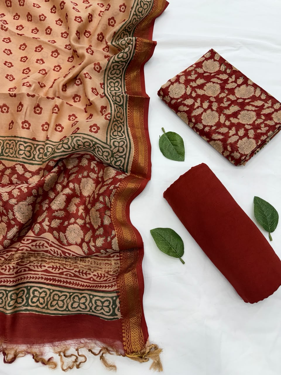 Full flat lay view of the Rust Red Bagru Hand Block Print Maheshwari Silk Suit Set, showcasing the folded cream dupatta with red block prints, a folded floral patterned kurta fabric, and a neatly rolled solid rust red bottom fabric styled with fresh green leaves.