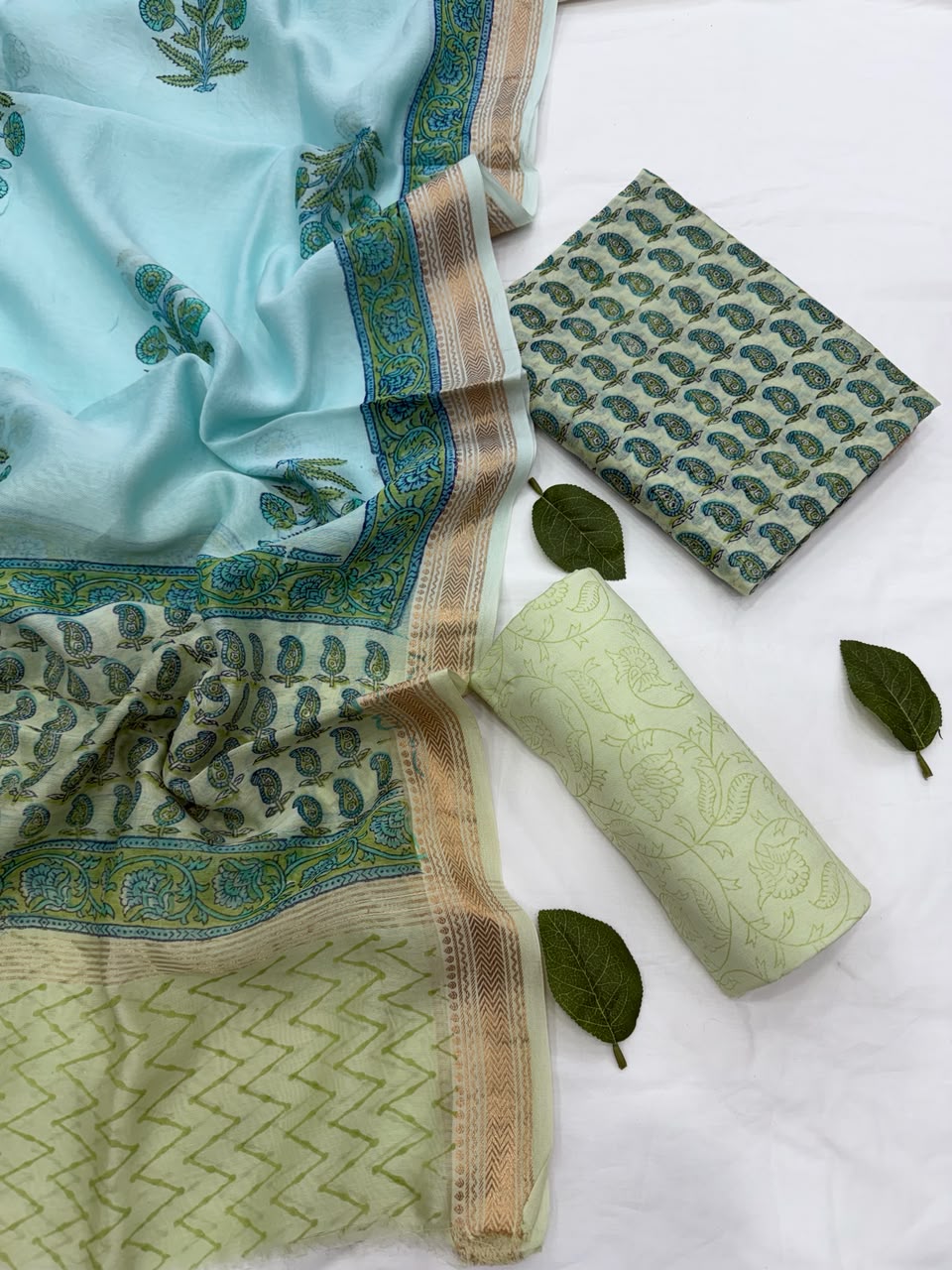 Full flat lay view of the Pista Green and Turquoise Sanganeri Buti Block Print Contrast Silk Suit Set, displaying the turquoise patterned dupatta with a golden Zari border, the matching folded kurta fabric, and a rolled pista green bottom fabric styled with fresh green leaves.