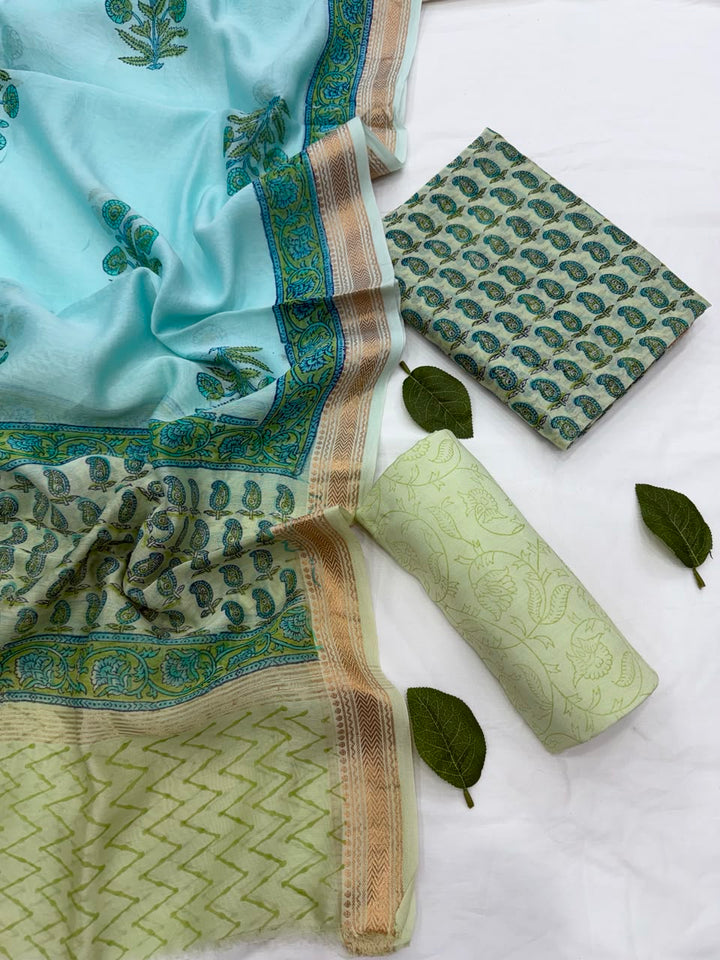 Full flat lay view of the Pista Green and Turquoise Sanganeri Buti Block Print Contrast Silk Suit Set, displaying the turquoise patterned dupatta with a golden Zari border, the matching folded kurta fabric, and a rolled pista green bottom fabric styled with fresh green leaves.
