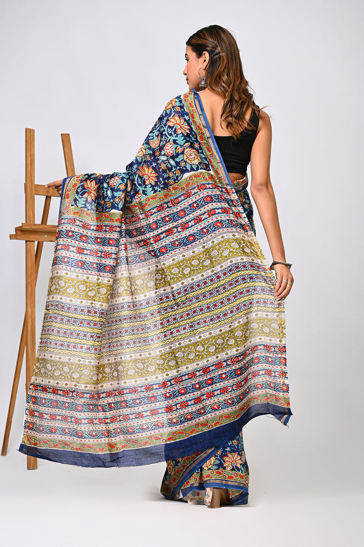 Back view of a woman modeling an exclusive blue Sanganeri print Chanderi Silk saree, showcasing the fluid drape of its intricate Kalamkari-inspired floral design and an elegant cream-patterned pallu next to a wooden easel.