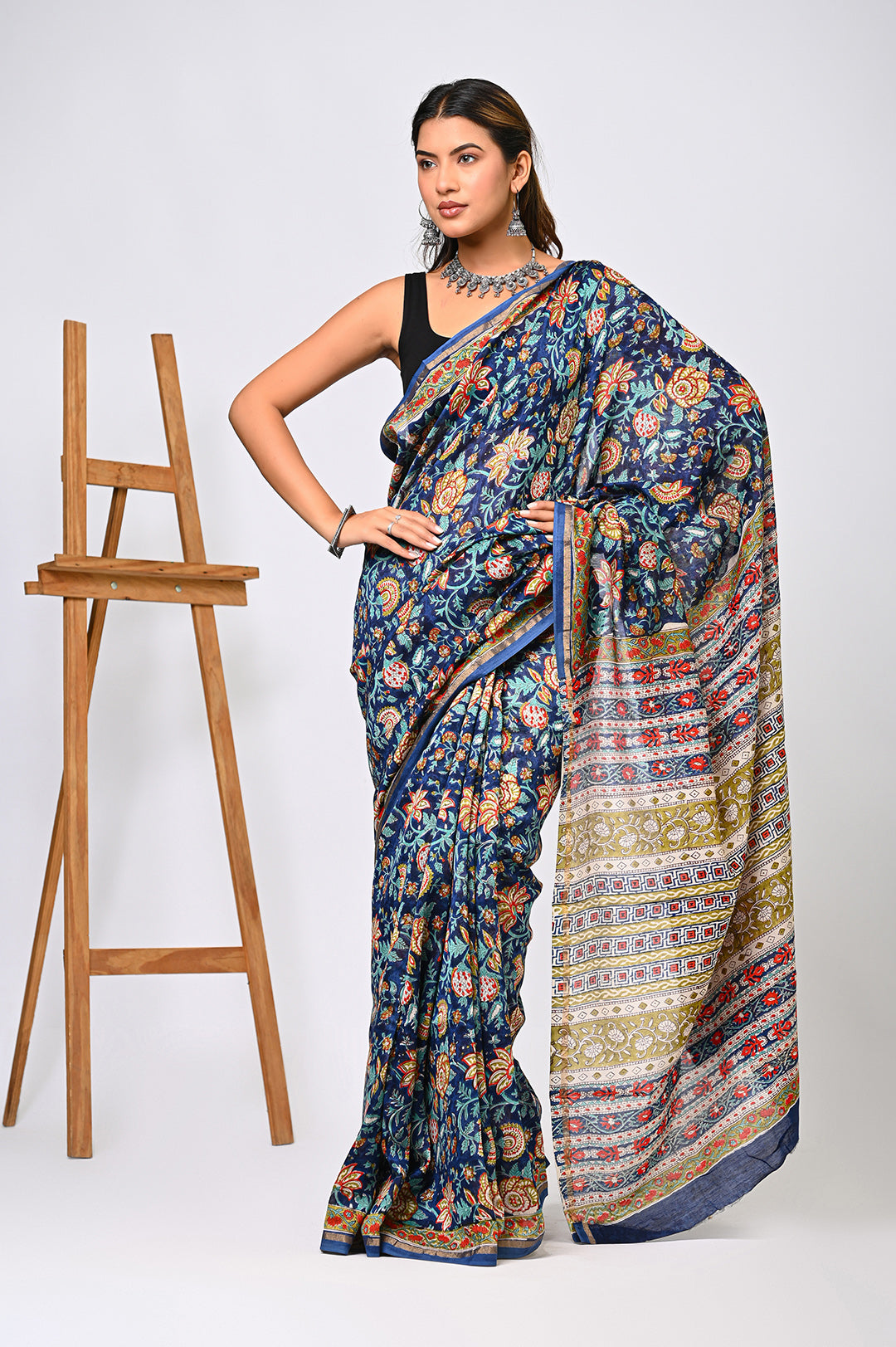 A woman modeling an exclusive blue Sanganeri print Chanderi Silk saree featuring an intricate Kalamkari-inspired floral design and an elegant cream-patterned pallu next to a wooden easel.