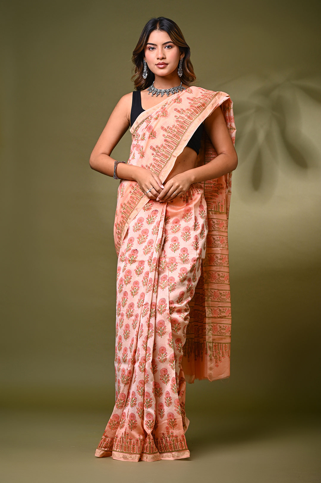 A woman modeling a soft peach Sanganeri print Chanderi Silk saree featuring delicate hand block printed floral butis, structured borders, and a matching patterned pallu against an olive green backdrop.