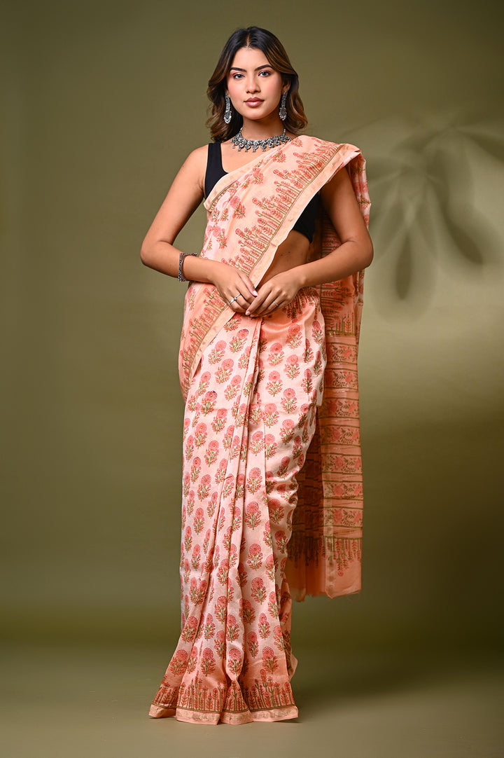 A woman modeling a soft peach Sanganeri print Chanderi Silk saree featuring delicate hand block printed floral butis, structured borders, and a matching patterned pallu against an olive green backdrop.