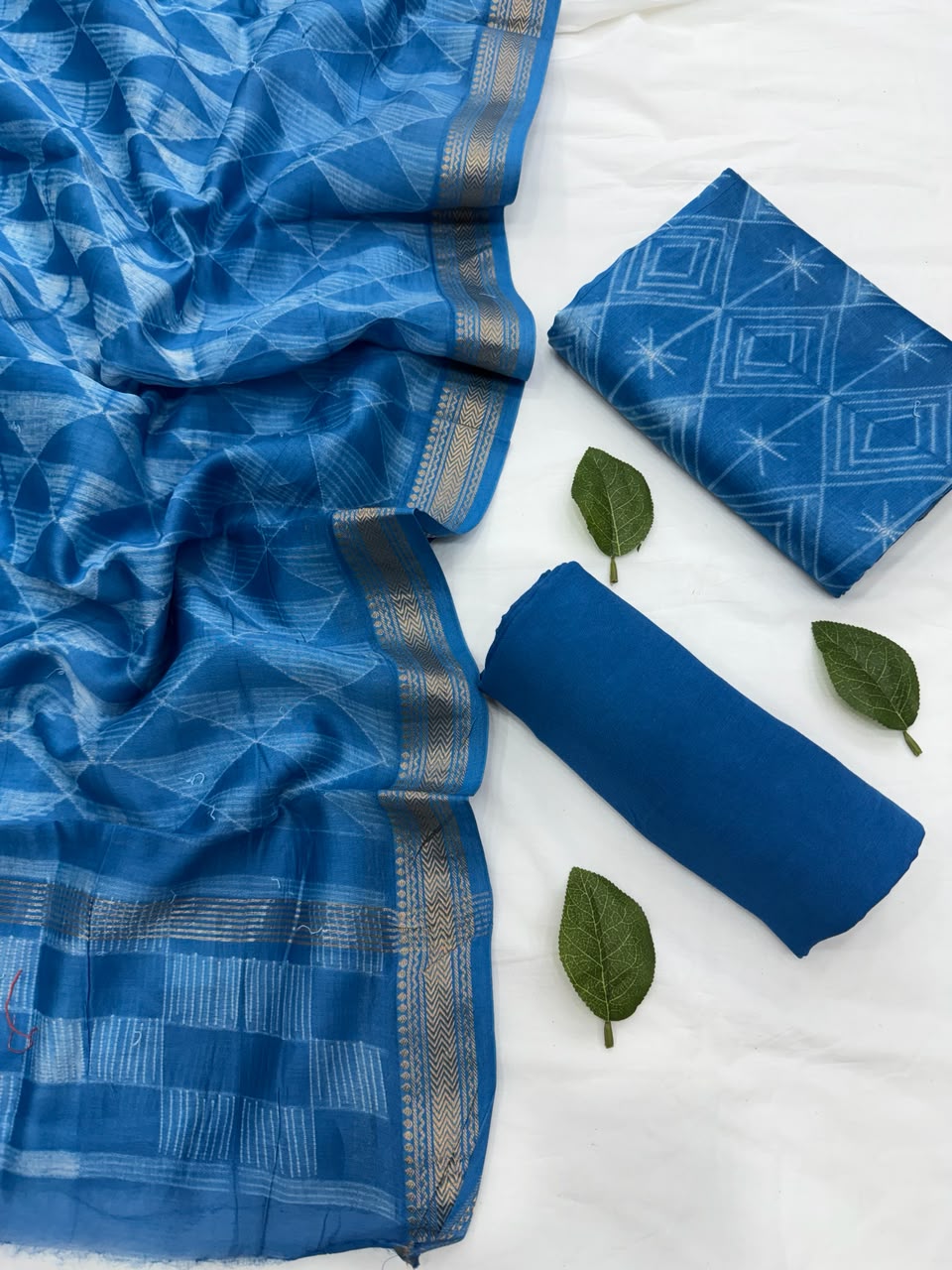 Full flat lay view of the Sky Blue Stitching Shibori Maheshwari Silk Suit Set, showcasing the shibori tie-dye patterned kurta fabric, a matching patterned dupatta with a golden handloom Zari border, and a neatly rolled solid sky blue bottom fabric decorated with green leaves.