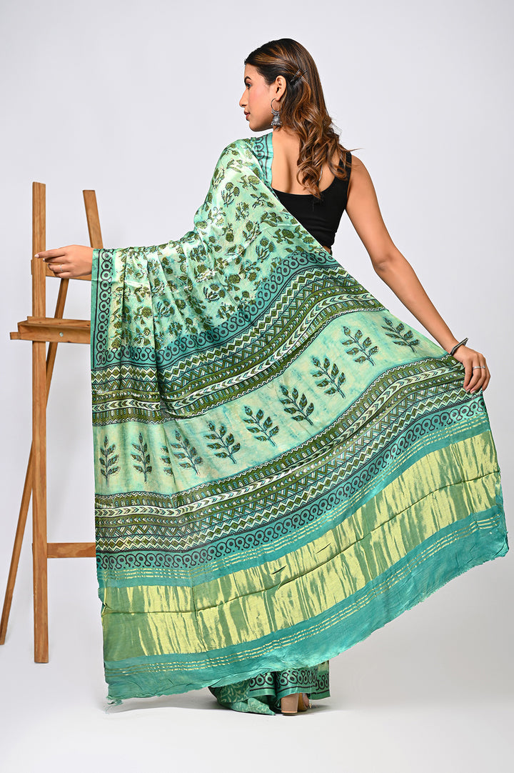 Back view of a woman modeling a natural green Vanaspati print Modal Silk saree, showcasing the fluid drape, intricate geometric block prints, and a traditional golden Lagdi Patta border next to a wooden easel.
