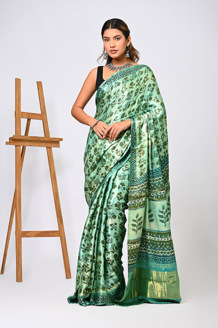 A woman modeling a natural green Vanaspati print Modal Silk saree featuring intricate geometric block prints, a traditional Lagdi Patta border, and an elegant patterned pallu next to a wooden easel.